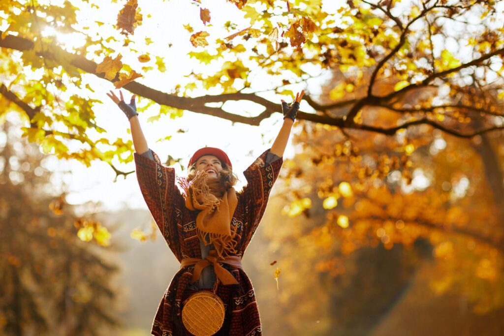 Eine Frau steht mit ausgebreiteten Armen unter herbstlichen Bäumen und genießt die warmen Sonnenstrahlen.