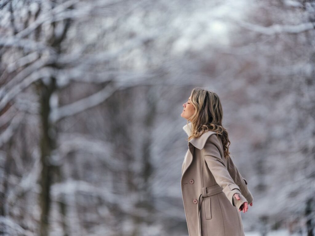 Eine Frau steht mit geschlossenen Augen in einer verschneiten Winterlandschaft und genießt die winterliche Stille.