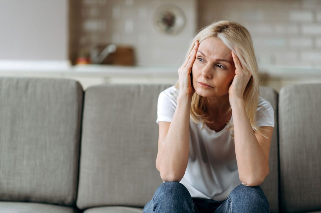 Eine erschöpfte Frau sitzt nachdenklich auf einem Sofa und stützt den Kopf in ihre Hände.