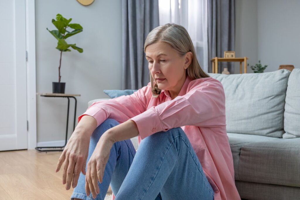 Eine nachdenkliche Frau sitzt auf dem Boden vor einem Sofa und blickt mit gesenktem Blick ins Leere.