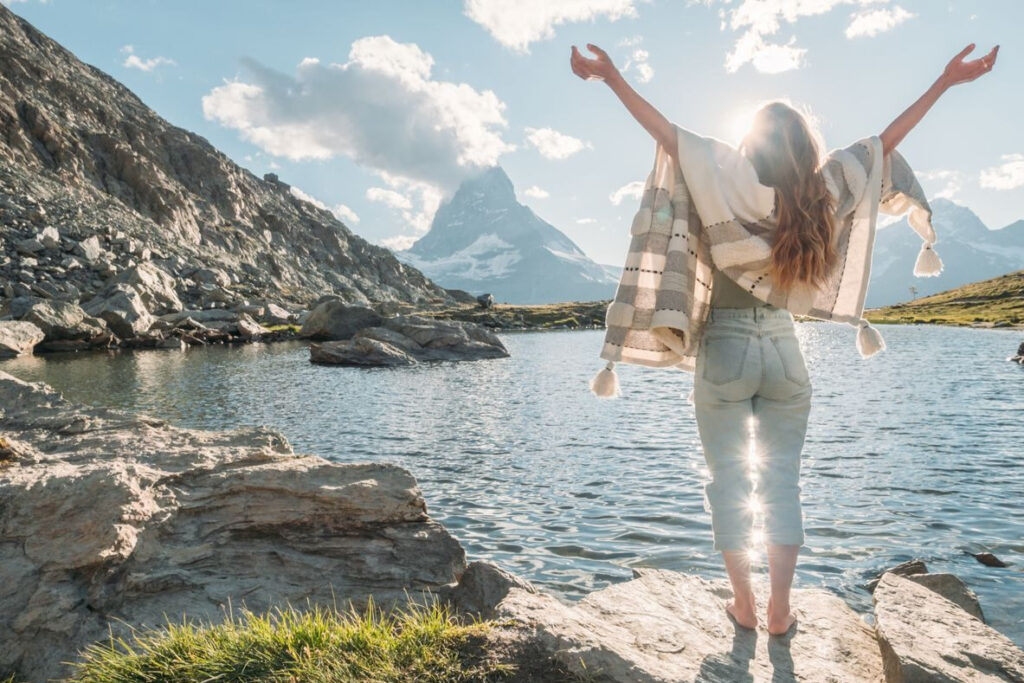 Eine Frau steht barfuß auf einem Felsen an einem Bergsee, breitet die Arme der Sonne entgegen aus und blickt auf eine imposante Berglandschaft.