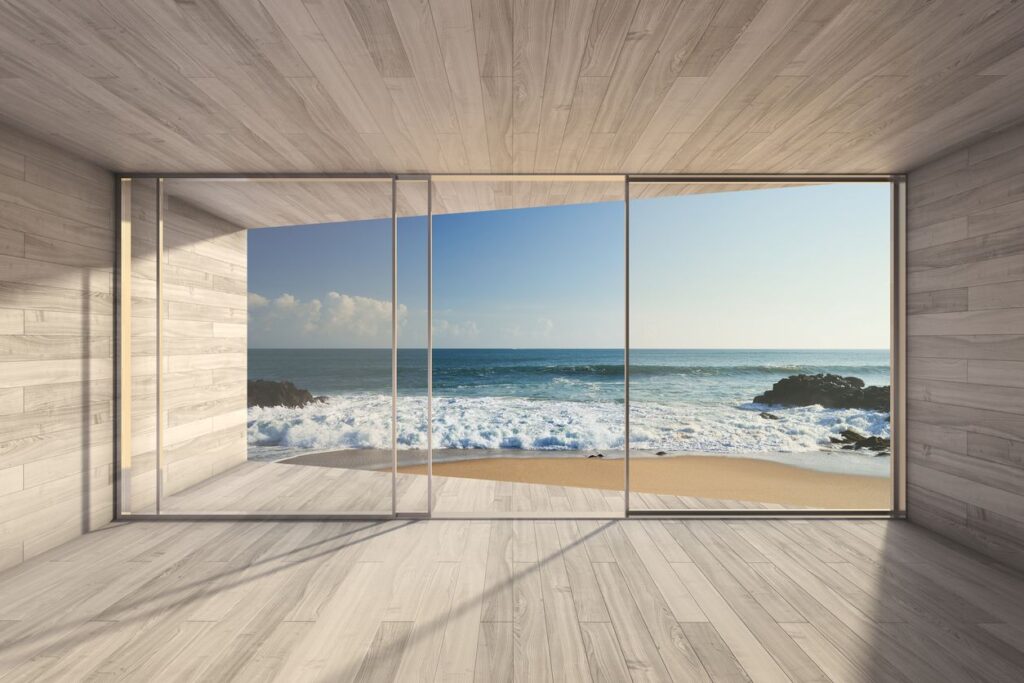 Ein heller, modern gestalteter Raum mit bodentiefen Glasfronten eröffnet den Blick auf einen Sandstrand, das Meer und die sanft brechenden Wellen.